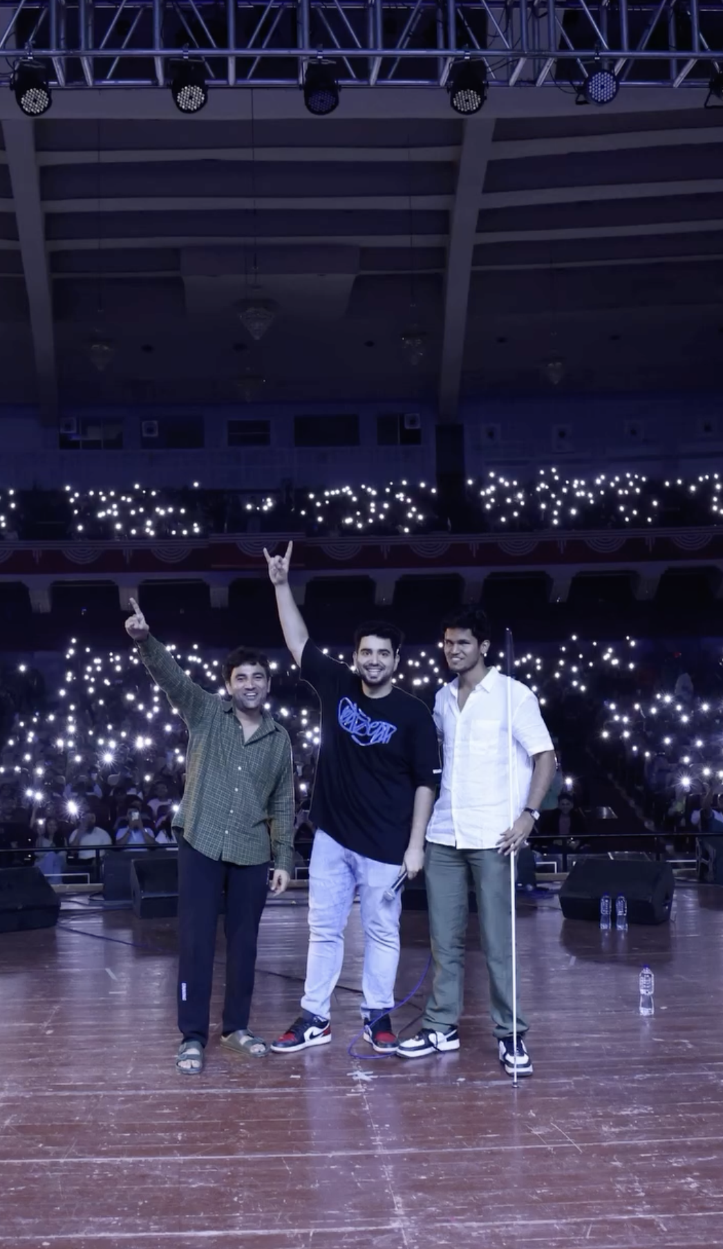 Left to right: Naman Arora, Samay Raina, and Bhavya Shah in different celebratory poses standing center stage in a large auditorium illuminated by thousands of phone flashlights